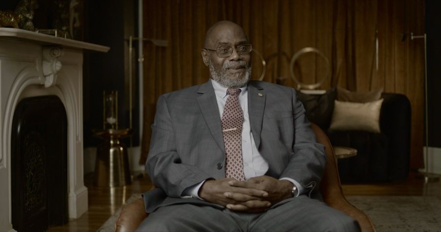 A lit documentary interview frame of a man seated in a wood-paneled room
