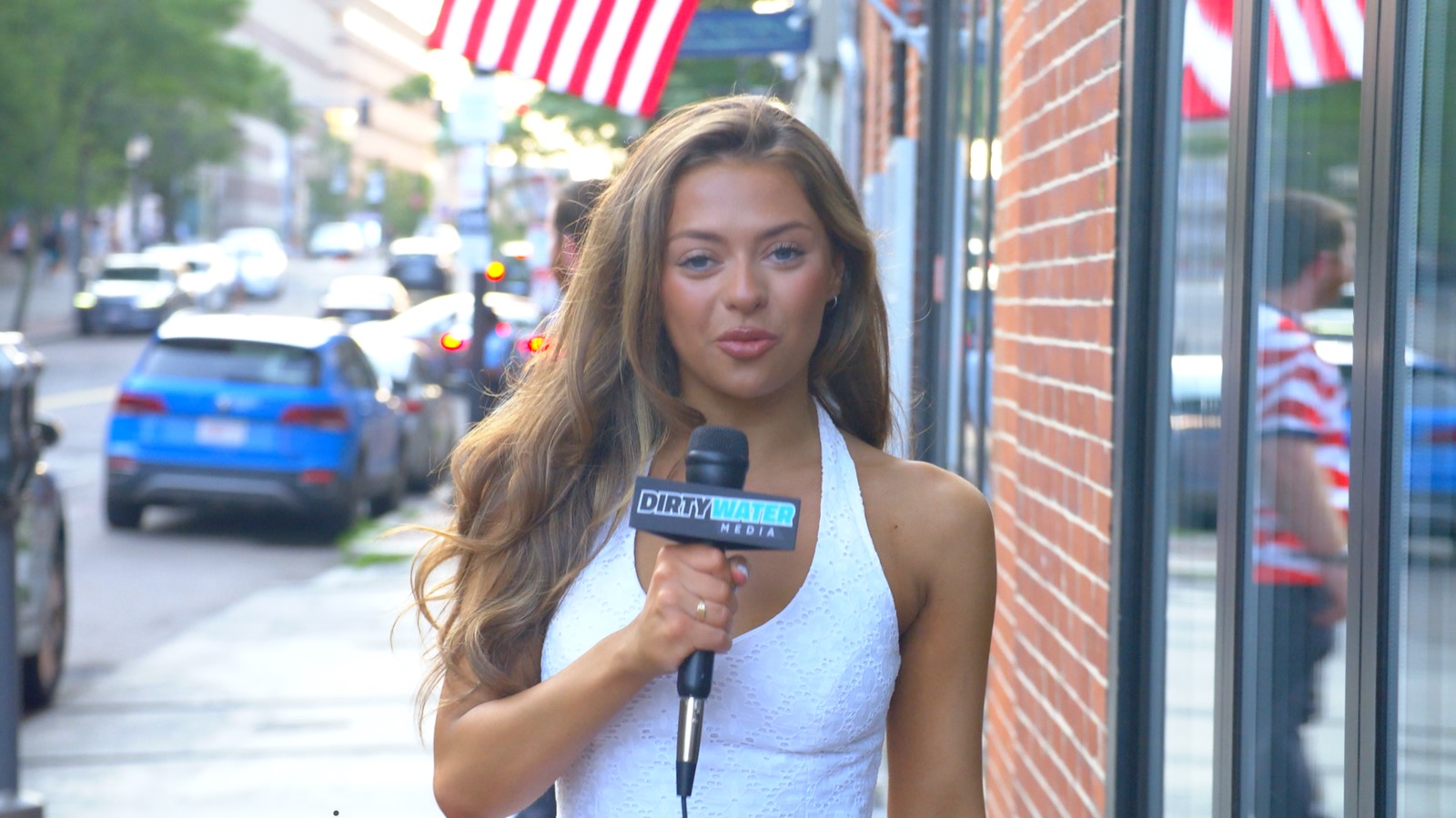 A Dirty Water Media reporter with a Dirty Water Media mic on a Boston sidewalk lined with American flags during the World Cup watch party at Clery's