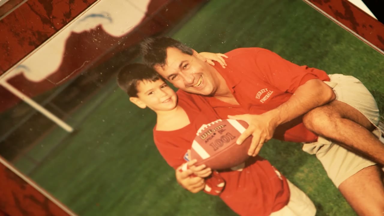 Archival photo of Coach DiBiaso in an Everett Football shirt holding a football with a young boy on the field, from the Coach Dib episode of Cardillo's World on NBC Sports Boston